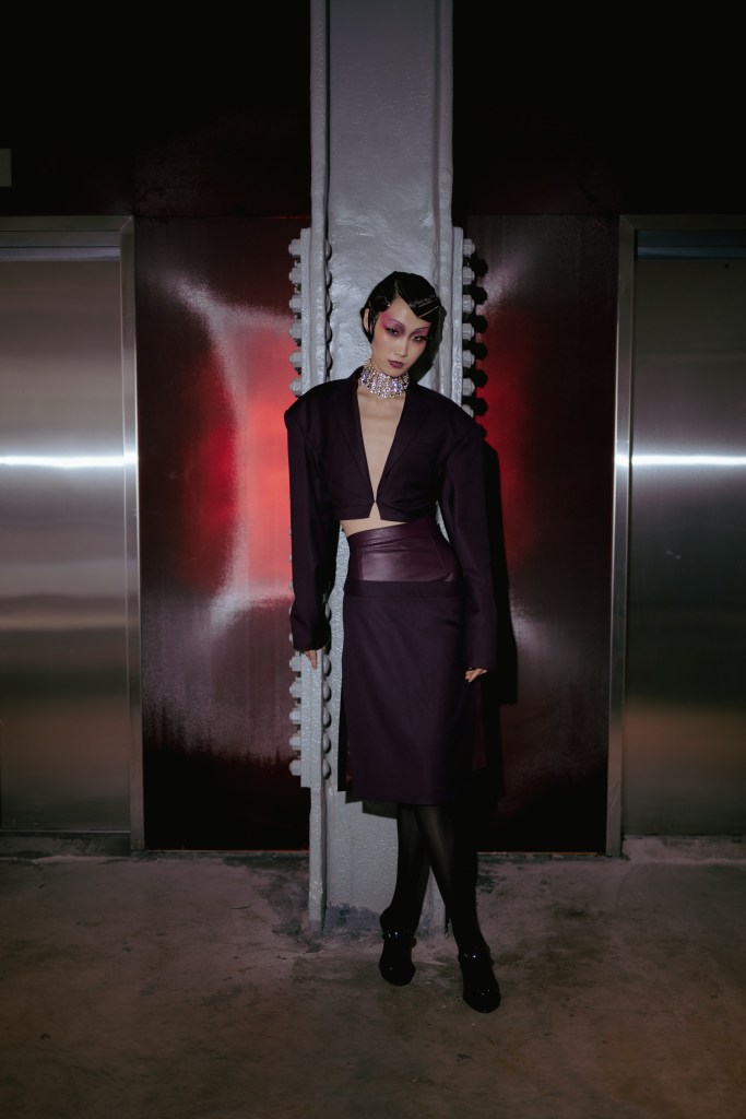 Shao New York Fall/Winter 2025 runway look featuring a cropped deep purple blazer with structured shoulders and a matching high-waisted leather-paneled midi skirt. The bold ensemble is accented by dramatic purple makeup, a sparkling statement choker, and sleek, sculpted hair. This look embodies a fusion of futuristic elegance and power dressing, highlighting the brand’s avant-garde approach to tailoring.