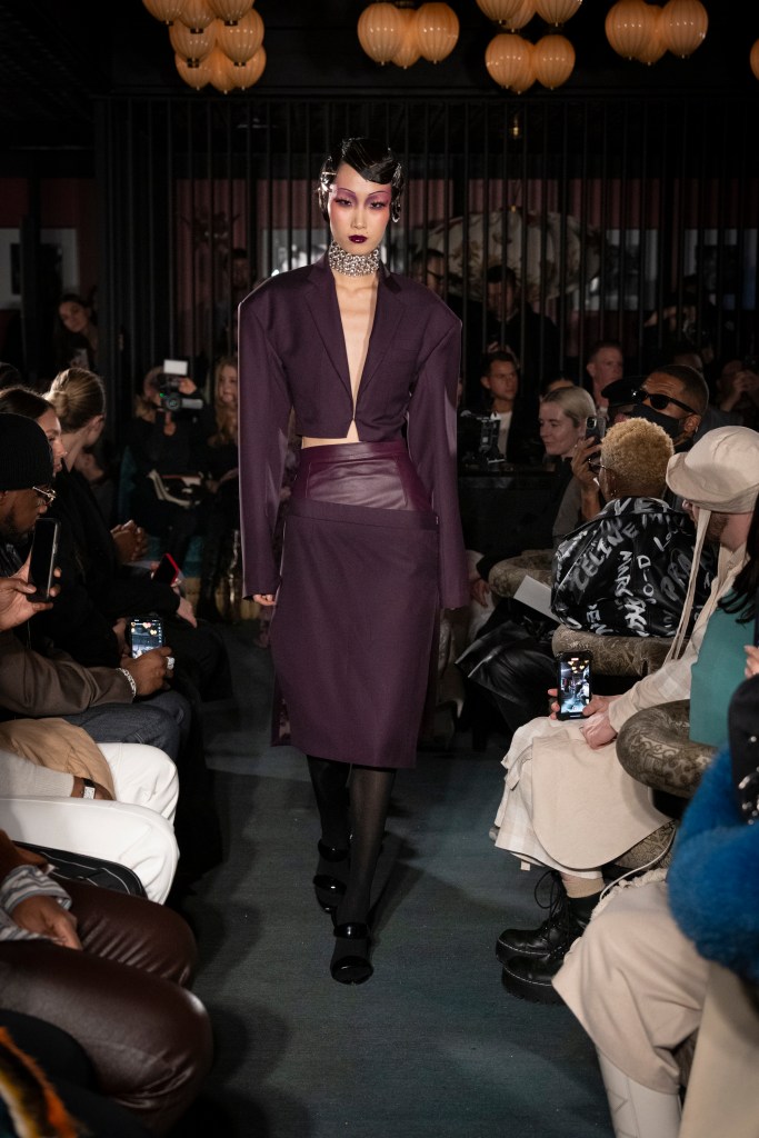 Shao New York Fall/Winter 2025 runway look featuring an oversized, deep purple blazer and matching wide-leg trousers. The ensemble is paired with a statement white shirt adorned with a bold graphic print. The look exemplifies modern tailoring with a contemporary, artistic twist, showcasing the brand's signature fusion of avant-garde silhouettes and streetwear influences.